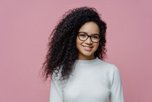 Headshot Of Lovely Curly Haired Woman With Healthy Skin And Toothy Smile, Dressed In White Jumper, Wears Transparent Glasses, Isolated Over Rosy Background. People, Face Expressions And Emotions