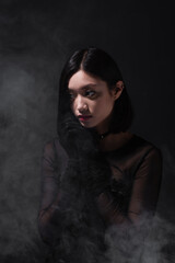 Wall Mural - portrait of young asian woman in gothic outfit and gloves looking away on grey with smoke.