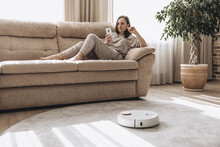 Woman Is Sitting On Couch And Operating White Robot Vacuum Cleaner Via Wi-fi. Girl Wearing Home Clothes Resting Relaxing Lying On Sofa With Smartphone. Smart Home