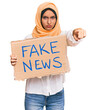 © Krakenimages.com - Young brunette arab woman holding fake news banner pointing with finger to the camera and to you, confident gesture looking serious