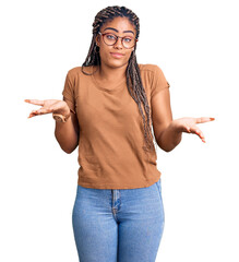 Wall Mural - Young african american woman with braids wearing casual clothes and glasses clueless and confused expression with arms and hands raised. doubt concept.