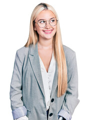 Beautiful blonde woman wearing business jacket and glasses looking away to side with smile on face, natural expression. laughing confident.