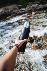 Wall Mural - Hand holds a thermos with tea, camping equipment for hiking, mountain hiking, water tank, vacuum bottle, mountain stream, bracelet on the arm