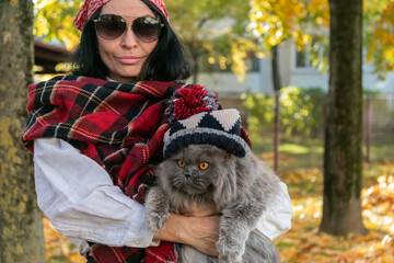 A girl in sunglasses with a scarf on her head holds a British cat in a hat with a pompom in her arms against the background of yellow autumn maple leaves. Walk with a cat.  Autumn mood. Close-up.