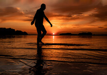 Silhouette At Sunset On Lake, Sea. Man Walking Along Seashore. Beautiful View Of Sundown With Orange Sky. Harmony, Calmness, Nature Beauty Concept