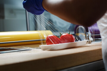 Unknown worker wraps in food transparent film tomatoes lying on white plastic tray.