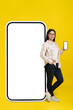 © Svyatoslav Lypynskyy - Looking at camera asian woman leaned on big smartphone with white screen showing phone in her hand wearing casual isolated on yellow background. Copy space on right, phone mock up