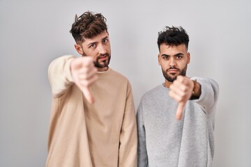 Wall Mural - Young homosexual couple standing over white background looking unhappy and angry showing rejection and negative with thumbs down gesture. bad expression.