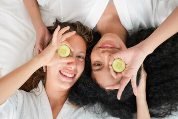 Two young attractive women in white bathrobes making facial mask holding slices of cucumber lying and chilling on bed posing to camera together in the bedroom, Women with beauty concept