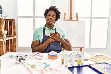 Wall Mural - Young african american man sitting on the table at art studio smiling with hands on chest with closed eyes and grateful gesture on face. health concept.