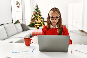 Canvas Print - Young caucasian girl sitting on the table working using laptop by christmas tree relaxed with serious expression on face. simple and natural looking at the camera.