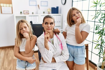 Sticker - Pediatrician woman working at the clinic with two little girls serious face thinking about question with hand on chin, thoughtful about confusing idea