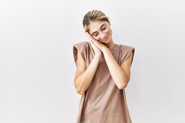 Wall Mural - Young blonde woman standing over isolated background sleeping tired dreaming and posing with hands together while smiling with closed eyes.