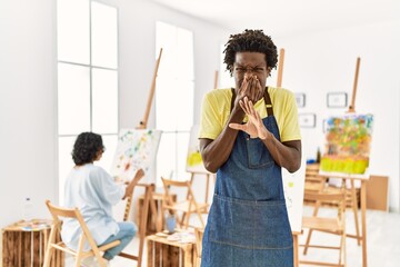 Poster - African young man standing at art studio smelling something stinky and disgusting, intolerable smell, holding breath with fingers on nose. bad smell