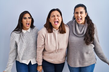 Wall Mural - Mother and two daughters standing over blue background angry and mad screaming frustrated and furious, shouting with anger. rage and aggressive concept.