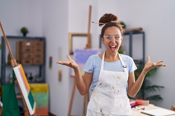 Wall Mural - Brunette woman painting at art studio celebrating victory with happy smile and winner expression with raised hands