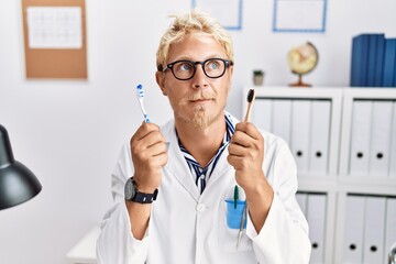 Poster - Young blond dentist man working at dentist clinic holding toothbrushes smiling looking to the side and staring away thinking.