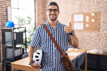 Canvas Print - Hispanic man with long hair working at the office holding bike helmet doing happy thumbs up gesture with hand. approving expression looking at the camera showing success.