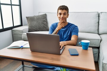 Sticker - Young handsome hispanic man using laptop sitting on the floor looking confident at the camera with smile with crossed arms and hand raised on chin. thinking positive.