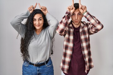 Poster - Young hispanic couple standing over white background doing funny gesture with finger over head as bull horns