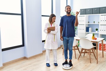 Poster - Dietitian at the clinic with client checking weight smiling happy pointing with hand and finger to the side