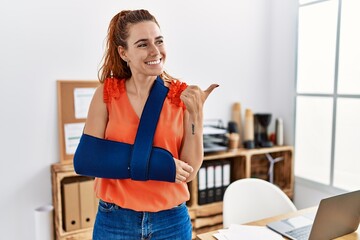 Poster - Young redhead woman wearing arm on sling at the office pointing thumb up to the side smiling happy with open mouth