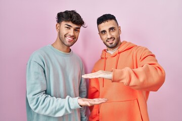 Wall Mural - Young hispanic gay couple standing over pink background gesturing with hands showing big and large size sign, measure symbol. smiling looking at the camera. measuring concept.