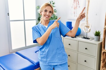 Poster - Beautiful blonde physiotherapist woman working at pain recovery clinic smiling and looking at the camera pointing with two hands and fingers to the side.