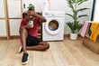 Young african american man using smartphone waiting for washing machine smiling and laughing with hand on face covering eyes for surprise. blind concept.