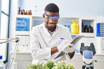 Canvas Print - Young african american man wearing scientist uniform writing on clipboard looking plant at laboratory