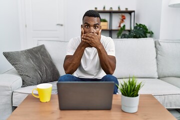 Wall Mural - Young african man using laptop at home shocked covering mouth with hands for mistake. secret concept.