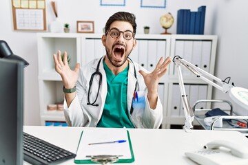 Sticker - Young man with beard wearing doctor uniform and stethoscope at the clinic celebrating crazy and amazed for success with arms raised and open eyes screaming excited. winner concept