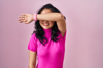 Poster - Young asian woman standing over pink background covering eyes with arm smiling cheerful and funny. blind concept.