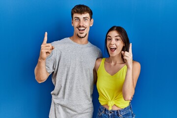 Young hispanic couple standing together over blue background pointing finger up with successful idea. exited and happy. number one.