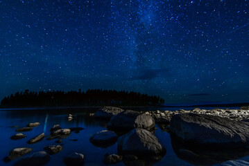 Wall Mural - night landscape with a starry sky and Milky way with many bright stars. Astrophotography with constellations and galaxies over water of a lake with rocks and a forest on background