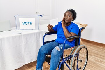 Poster - Young african woman sitting on wheelchair voting putting envelop in ballot box hand on mouth telling secret rumor, whispering malicious talk conversation