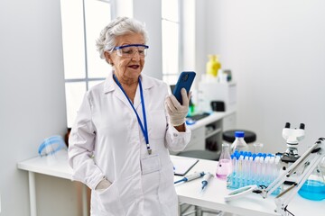 Wall Mural - Senior grey-haired woman wearing scientist uniform using smartphone at laboratory