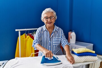 Wall Mural - Senior grey-haired woman smiling confident ironing clothes at laundry room