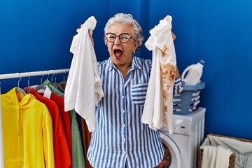 Wall Mural - Senior woman with grey hair holding clean white t shirt and t shirt with dirty stain angry and mad screaming frustrated and furious, shouting with anger. rage and aggressive concept.