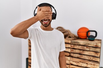 Canvas Print - African american man listening to music using headphones at the gym smiling and laughing with hand on face covering eyes for surprise. blind concept.