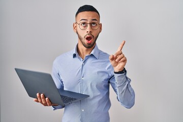 Poster - Young hispanic man working using computer laptop amazed and surprised looking up and pointing with fingers and raised arms.