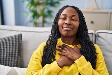 Wall Mural - African american woman sitting on sofa with hands on heart at home