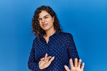 Poster - Young brunette woman with curly hair wearing casual clothes over blue background disgusted expression, displeased and fearful doing disgust face because aversion reaction. with hands raised