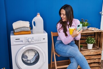 Wall Mural - Young hispanic woman using smartphone and drinking coffee waiting for washing machine at laundry room