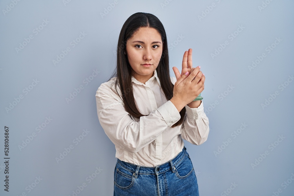 Young latin woman standing over blue background holding symbolic gun with hand gesture, playing killing shooting weapons, angry face
