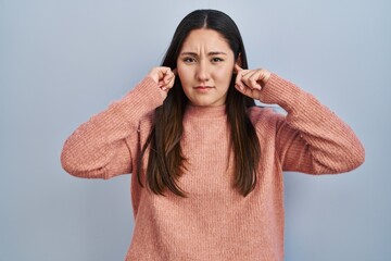 Sticker - Young latin woman standing over blue background covering ears with fingers with annoyed expression for the noise of loud music. deaf concept.