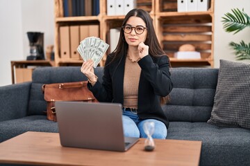 Poster - Young woman working at consultation office holding money serious face thinking about question with hand on chin, thoughtful about confusing idea