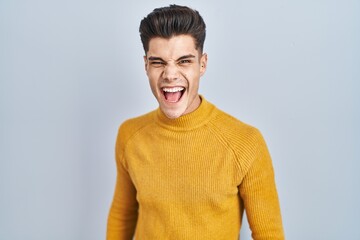 Sticker - Young hispanic man standing over blue background angry and mad screaming frustrated and furious, shouting with anger. rage and aggressive concept.