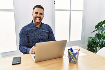 Poster - Young hispanic man with beard working at the office with laptop sticking tongue out happy with funny expression. emotion concept.