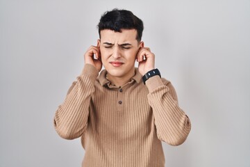 Sticker - Non binary person standing over isolated background covering ears with fingers with annoyed expression for the noise of loud music. deaf concept.